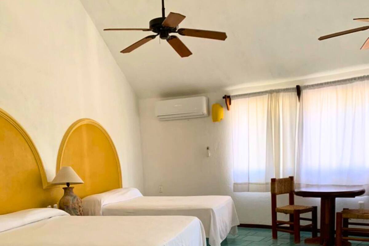A hotel room with two beds, a ceiling fan, an air conditioning unit, a table with chairs, and a window with curtains.