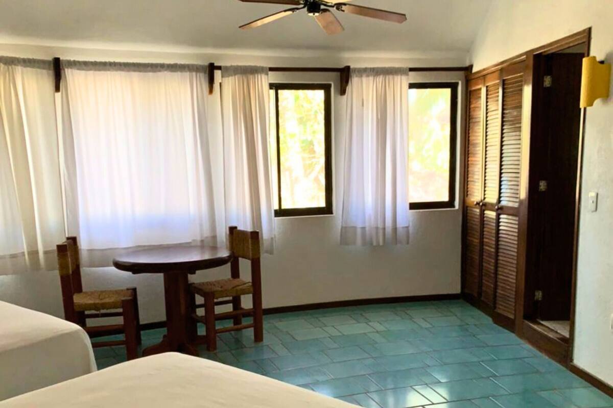 A room with two beds, a ceiling fan, a window with curtains, a table with chairs, and a door with louvered shutters.