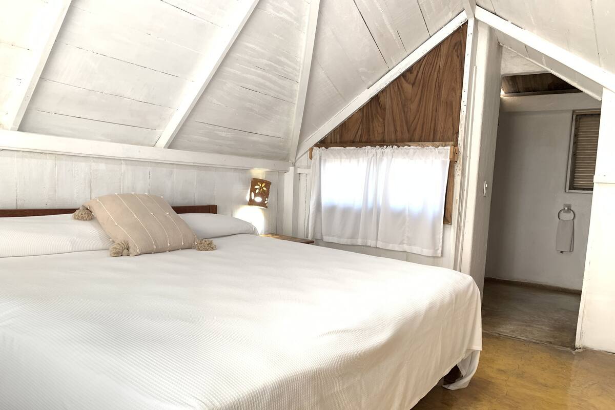 A bedroom with a sloped ceiling, a bed with white bedding, a nightstand with a lamp, and a window with curtains.