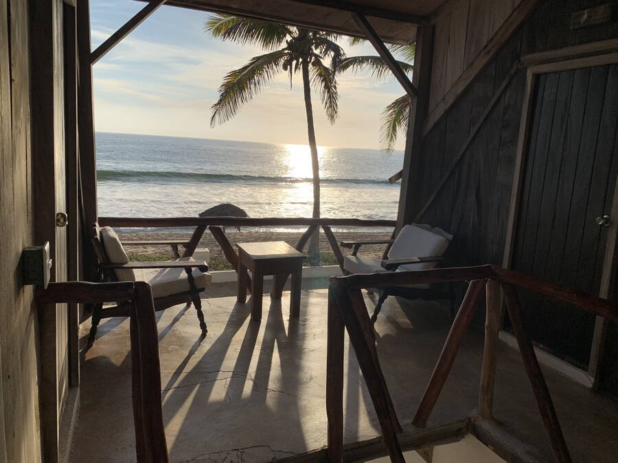 A wooden deck with a view of the ocean and palm trees. There are two chairs and a table on the deck.