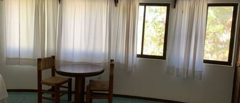 A room with a round table and four chairs, large windows with white curtains, and a ceiling fan.