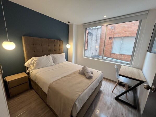 A bedroom with a large bed, bedside tables, a window with a view of a brick building, and a small desk with a chair.