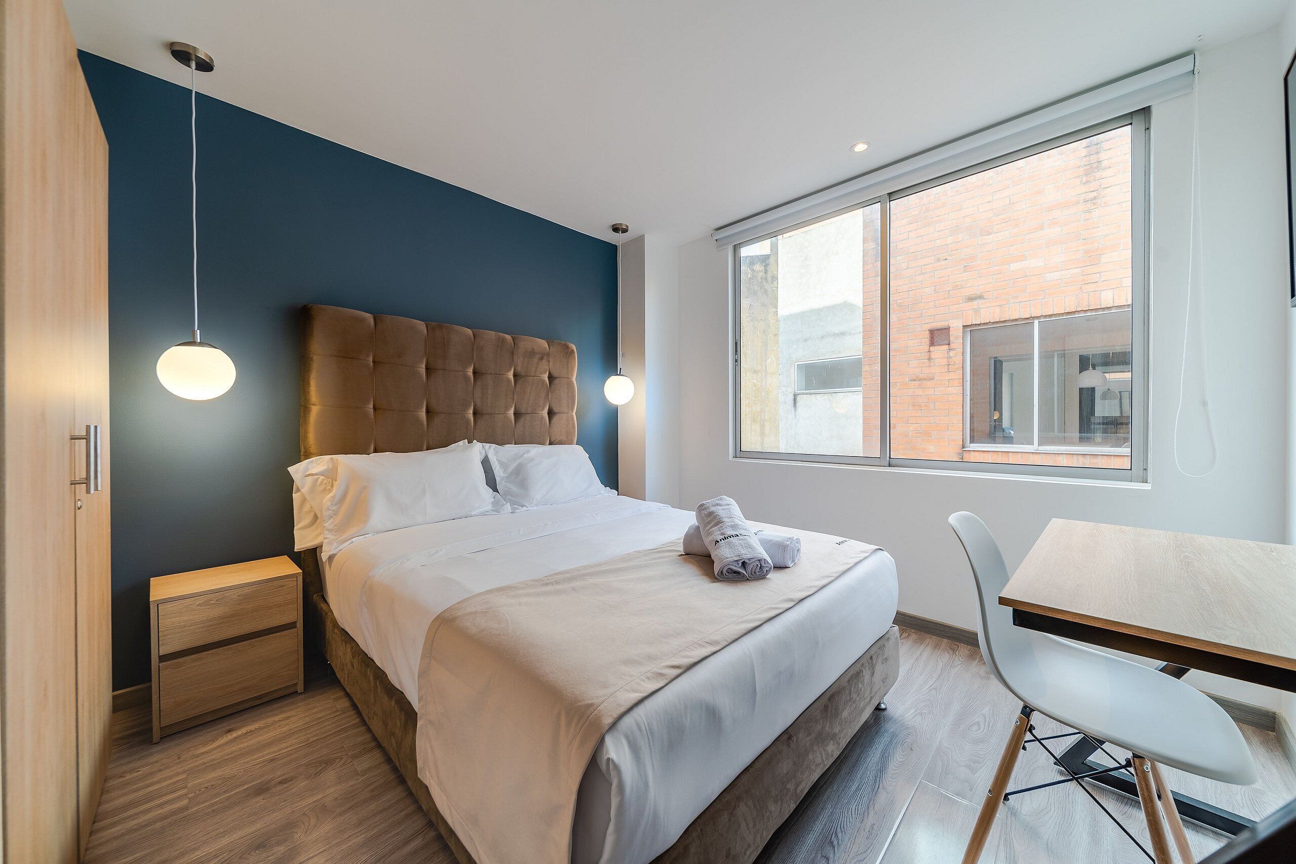 A modern bedroom with a large bed, bedside tables, a desk, and a window with a view of a brick building.