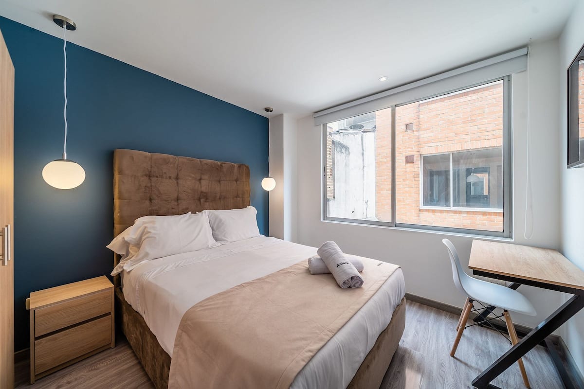 Une chambre moderne avec un grand lit, une table de chevet en bois, un bureau et une vue sur un bâtiment en briques, visible à travers une fenêtre.