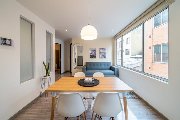 A modern dining area with a wooden table, white chairs, a blue sofa, and a large window offering a view of the outside.
