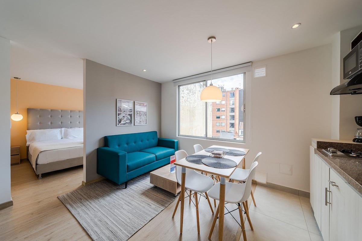 Une pièce moderne et bien éclairée, comprenant un lit, un canapé bleu, une table à manger avec des chaises et un coin cuisine.