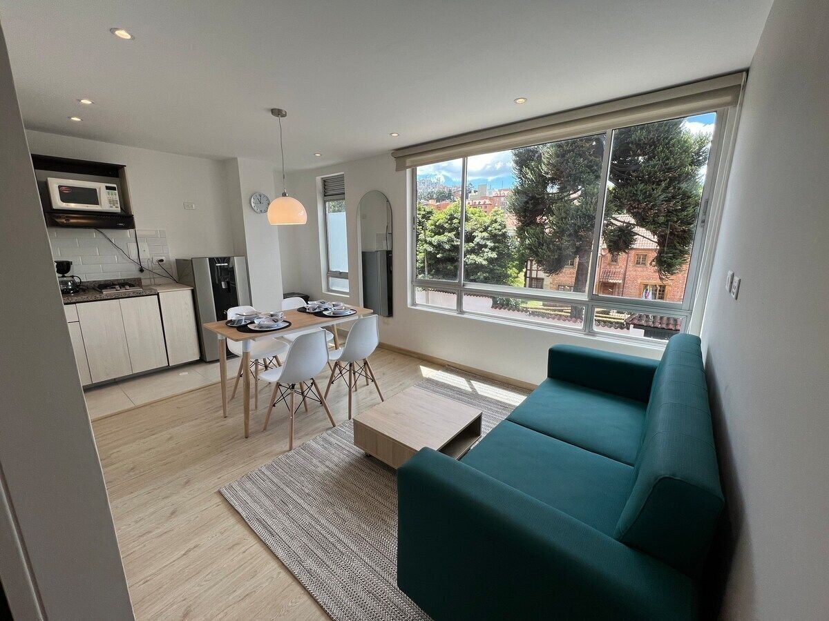 A modern living room with a teal sofa, a dining area with a table and chairs, and a kitchen with a microwave.