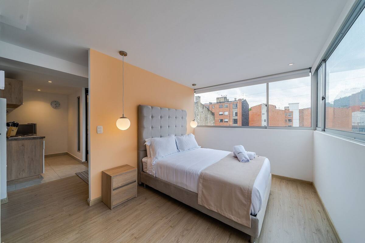 Une chambre moderne avec un grand lit, des tables de chevet, une armoire et une vue sur les bâtiments par une grande fenêtre.