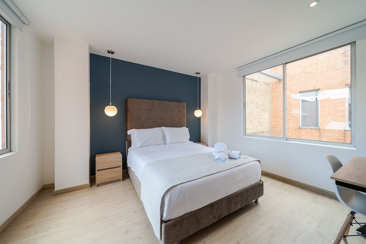 Une chambre moderne avec un grand lit, des tables de chevet, un bureau et une fenêtre donnant sur un immeuble en briques.