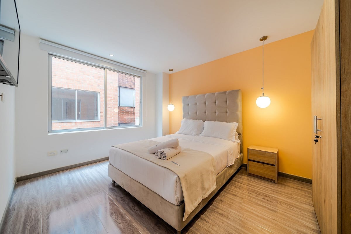 Une chambre moderne avec un grand lit, une table de chevet et une fenêtre donnant sur un immeuble en briques.