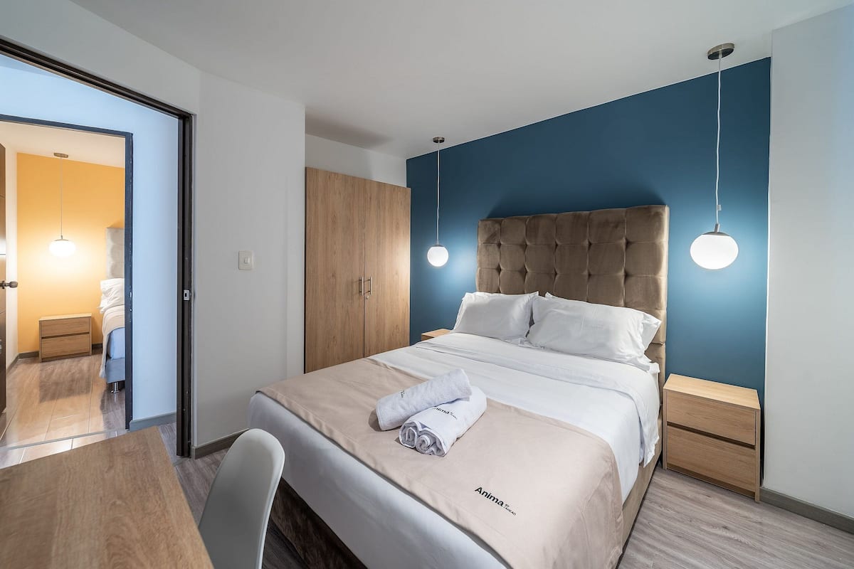 Un lit bien fait, avec une couette blanche et une serviette pliée, une table de chevet en bois avec un tiroir, et un mur d’accent bleu.
