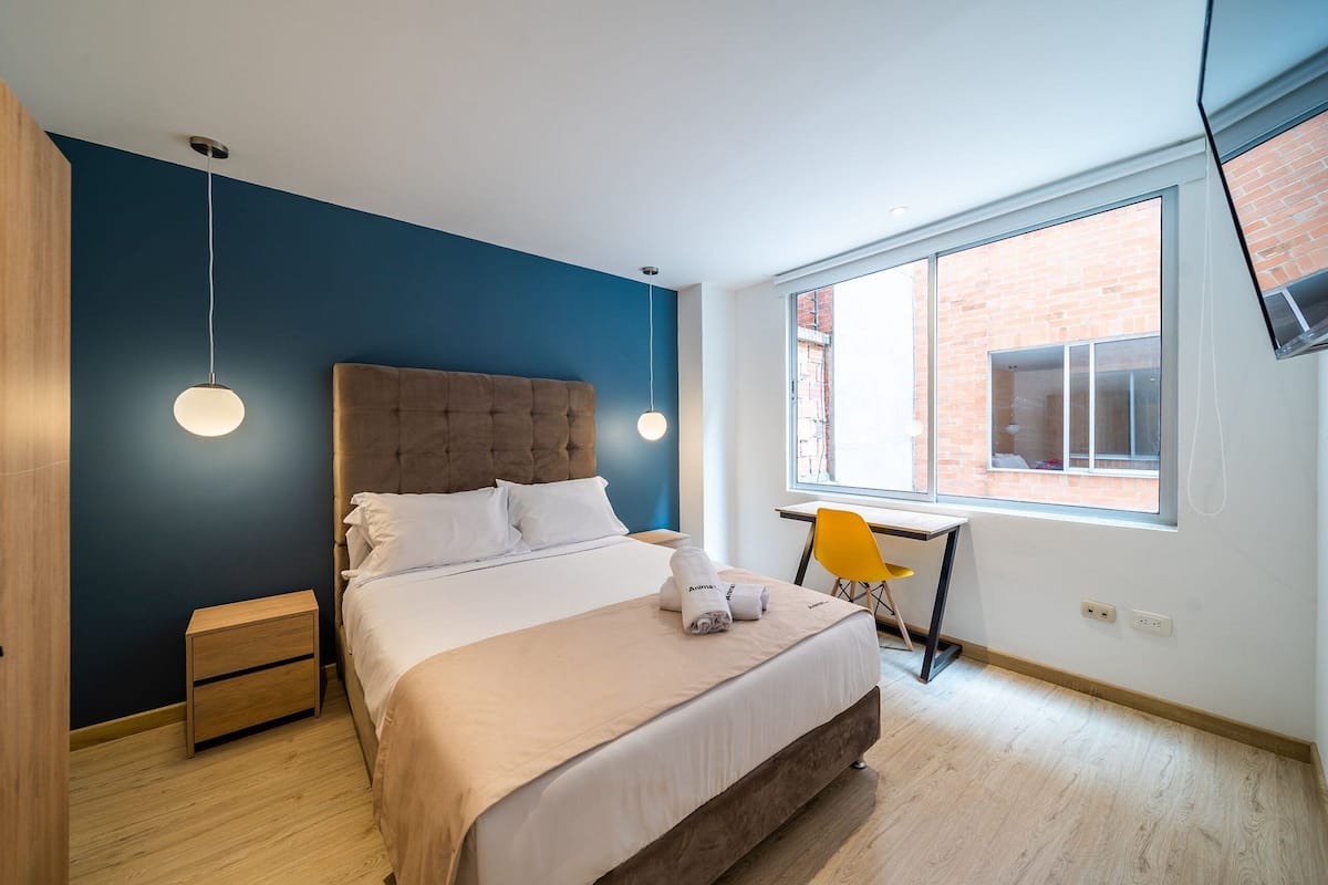 Une chambre moderne avec un grand lit, des tables de chevet, un bureau et une fenêtre donnant sur un immeuble en briques.