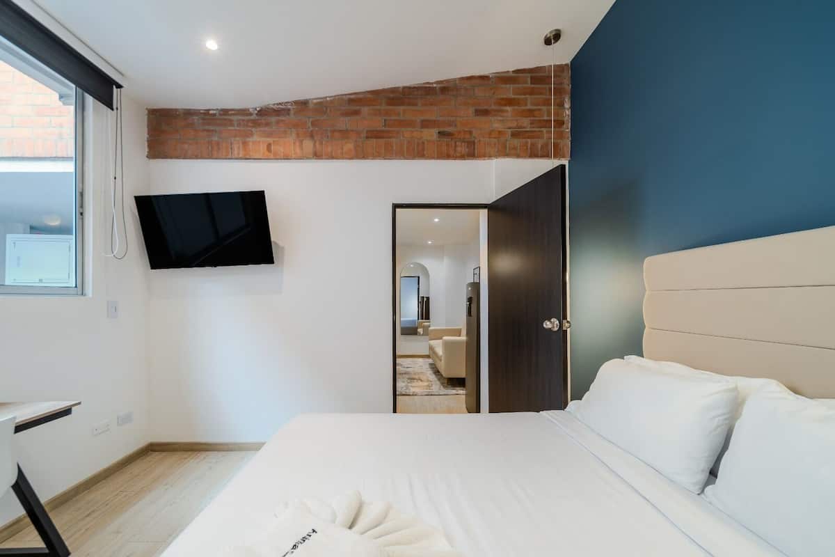 Une chambre avec un mur d’accent en briques, un téléviseur à écran plat fixé au-dessus du lit, un plancher en bois et une porte donnant sur une salle de bain.