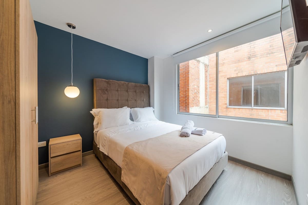 Une chambre moderne avec un grand lit, une table de chevet, une lampe suspendue et une vue sur un bâtiment en briques, visible à travers une fenêtre.