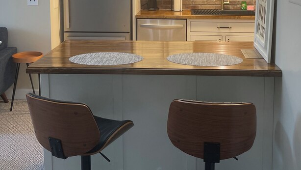 A compact kitchen with a refrigerator, two bar stools, a wooden countertop, and a brick backsplash.