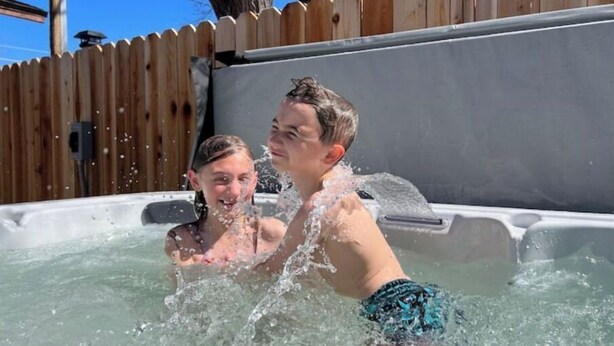 A hot tub with two people in it, surrounded by a wooden fence and a clear blue sky. - 2 min from rt 66- hot tub-firepit-bocceball-bbq (Williams)