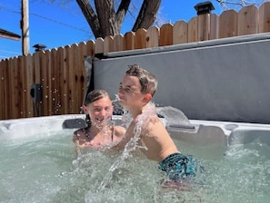 A hot tub with two people in it, surrounded by a wooden fence and a clear blue sky. - 2 min from rt 66- hot tub-firepit-bocceball-bbq (Williams)