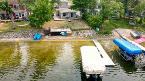 A lake with a dock and a covered boat. There are houses and trees in the background. - Lake Front, Pontoon, Bunk House (Rochert)