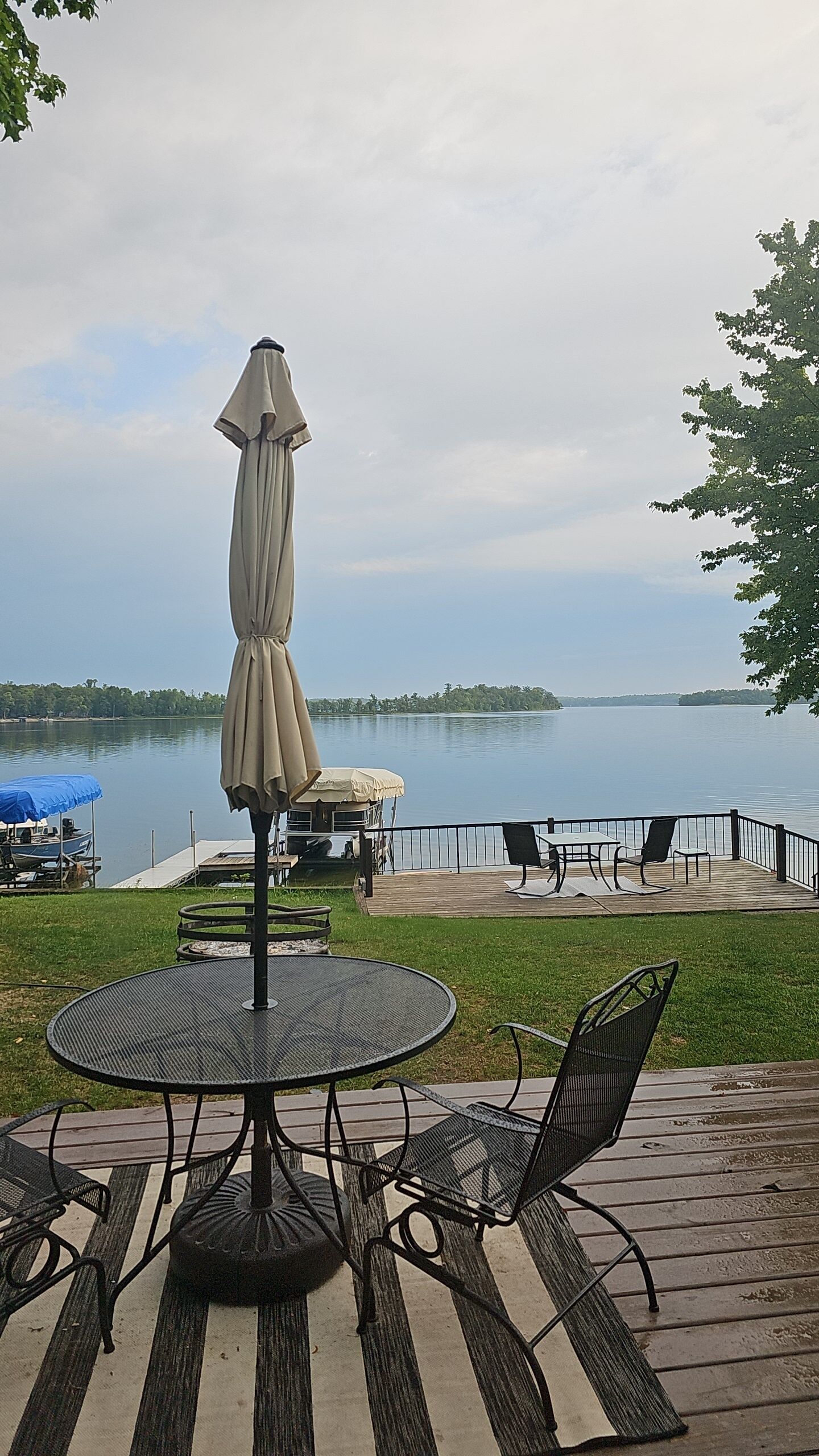 An outdoor patio with a round table and chairs, a beige umbrella, and a view of a lake with a dock and boats in the background.