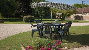 An outdoor dining area with a table and chairs under a plaid umbrella, surrounded by a garden with flowers and a hedge. - Les Rosiers - ONJON - Gîte (ONJON)