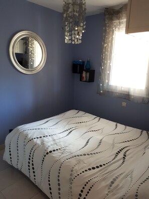 A bedroom with a bed, a mirror, a shelf with books and a vase, and a window with curtains. - Apartment with a breathtaking ocean view (Banyuls-sur-Mer)