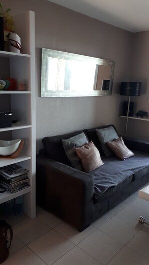 A living room with a dark grey sofa, two cushions, a white shelf with various items, a large mirror, and a floor lamp. - Apartment with breathtaking sea view (Banyuls-sur-Mer)