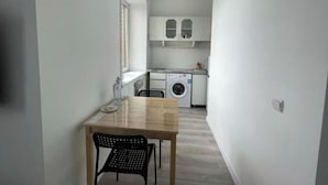 A narrow hallway leading to a kitchen with a washing machine, a table with chairs, and a window providing natural light.