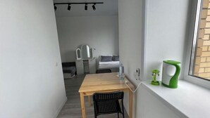 A narrow room with a wooden table and a black chair, a small window with a green kettle, and a bed with a white headboard in the background.