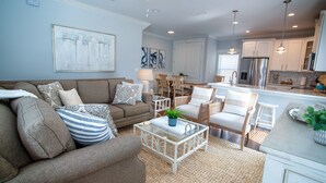 A living room with a brown sofa, white armchairs, a glass coffee table, and a dining area with wooden chairs. - Townhome Steps to the Beach-Hot Tub, Private Yard (Jacksonville Beach)
