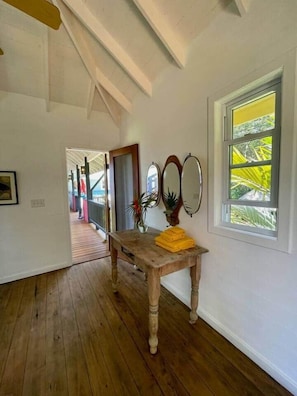 A hallway with wooden flooring, a wooden table with yellow towels, a mirror, and a window with a view of greenery.