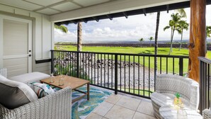 A balcony with wicker furniture, a view of a golf course, and a wooden table with a vase. - Luxury Living with Pool Access (Kamuela)