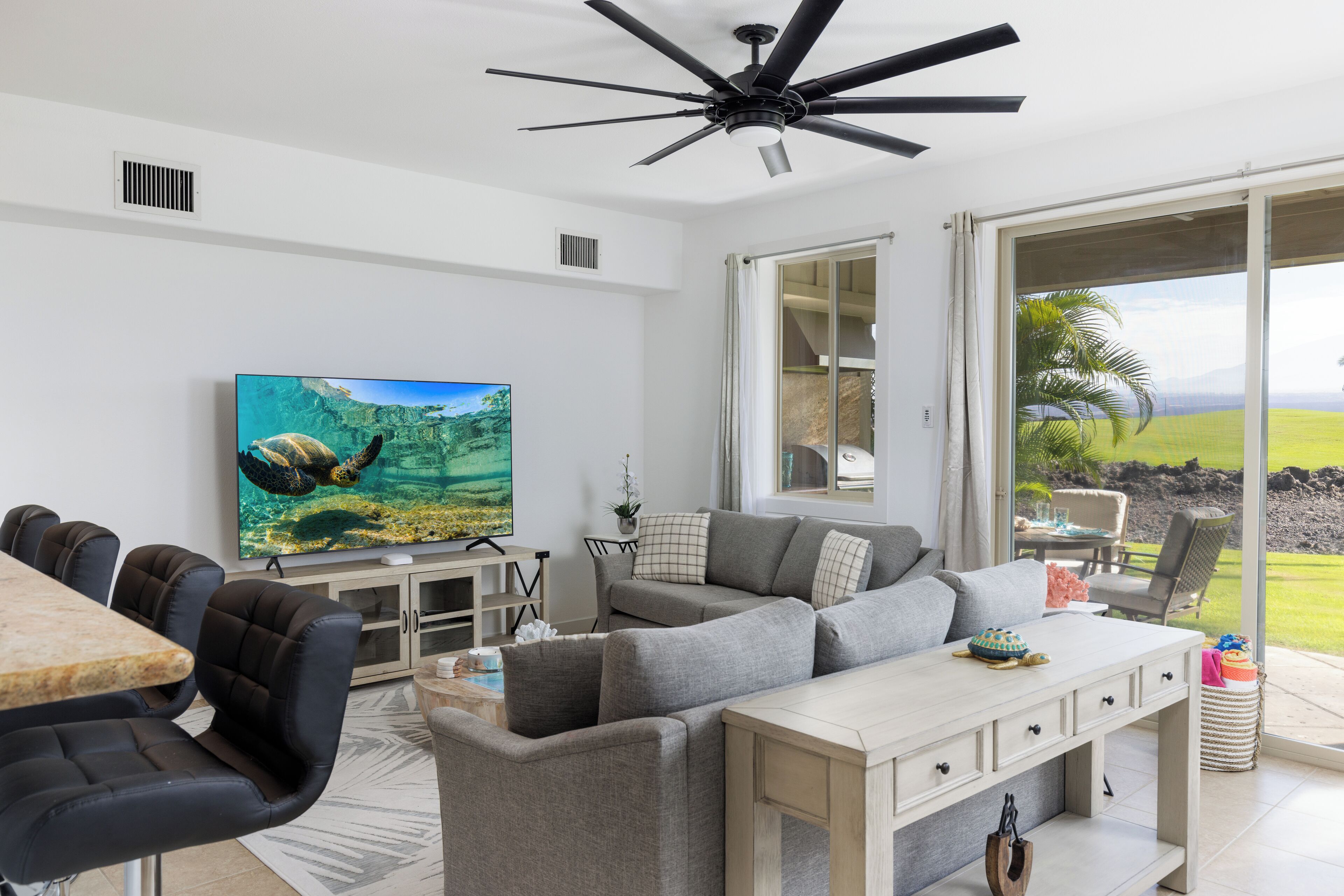 Ein modernes Wohnzimmer mit einem großen Flachbildfernseher, einem Deckenventilator und einer Schiebetür zu einem Balkon mit Landschaftsblick.