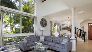 A spacious living room with a grey sectional sofa, a glass coffee table, and a view of palm trees through large windows. - Luxury Living with Pool Access (Kamuela)
