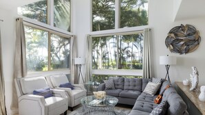 A spacious living room with a grey sofa, two armchairs, a glass coffee table, and large windows offering a view of palm trees. - Luxury Living with Pool Access (Kamuela)
