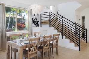 A dining area with a wooden table set for four, a staircase with black railings, and a large window with a view of trees. - Luxury Living with Pool Access (Kamuela)
