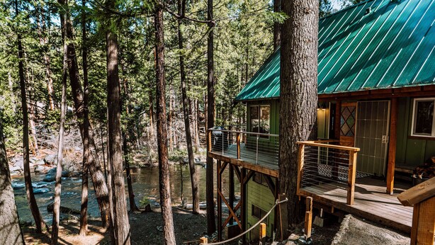 A wooden cabin with a green roof, a deck, and a view of a river surrounded by trees.
