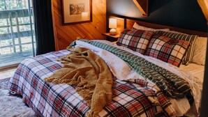 A wooden cabin bedroom with a bed, a nightstand, a lamp, and a window with a view of trees.