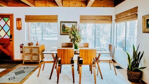 A dining area with wooden ceiling beams, a white table with four chairs, a vase with flowers, and a large window with blinds.