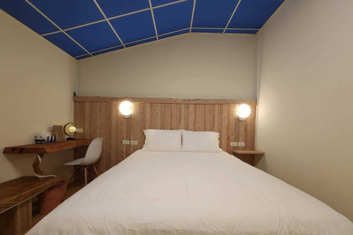 Ein Schlafzimmer mit einem hölzernen Kopfende, einem Bett mit weißer Bettwäsche, einem Schreibtisch mit Stuhl und zwei Nachttischlampen.