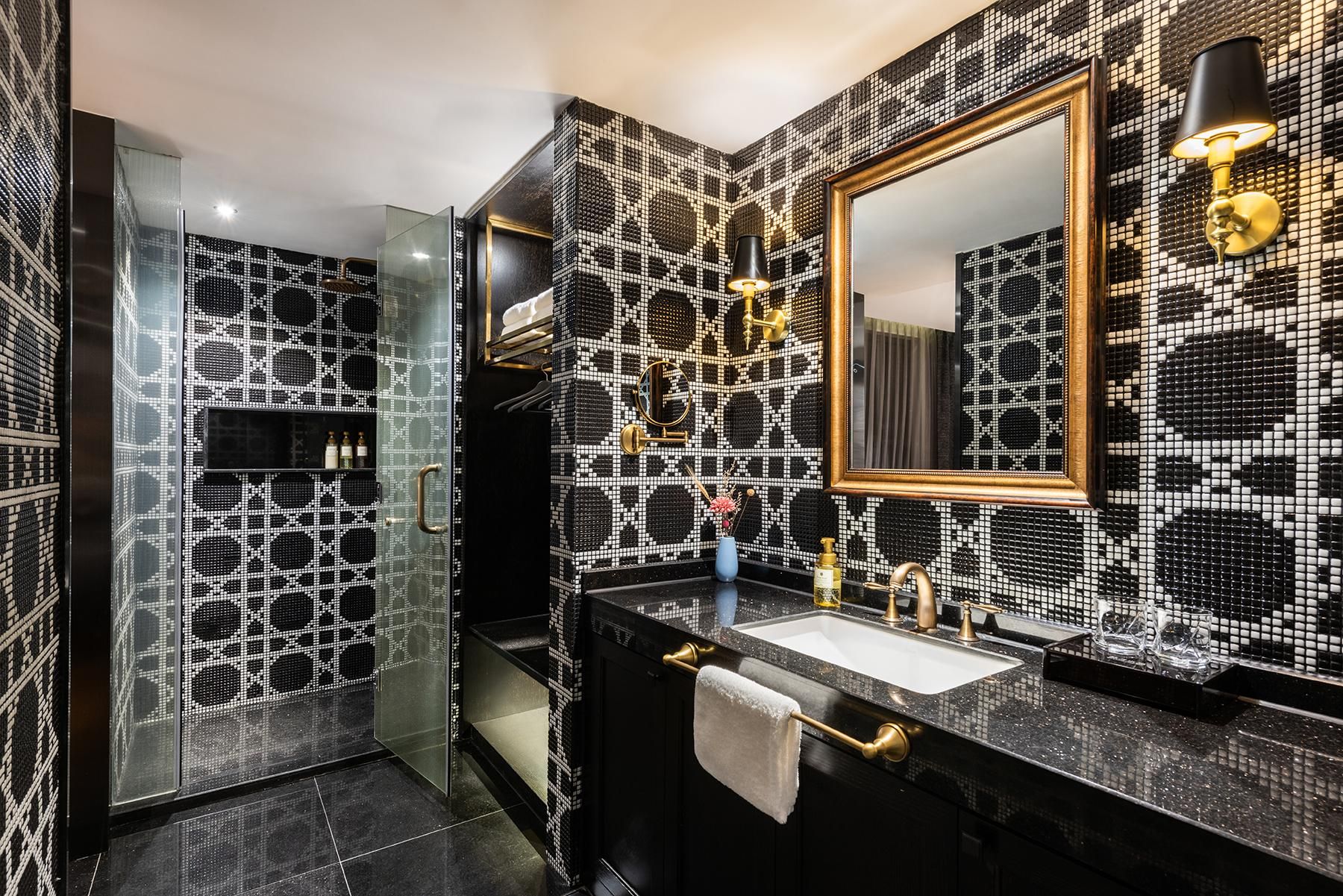 Une salle de bain avec un mur recouvert de mosaïques noires et blanches, un miroir à cadre doré, un plan de toilette noir et un robinet doré.