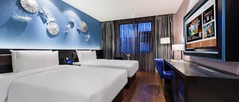 A hotel room with two beds, a desk, a television, and a large window with curtains.