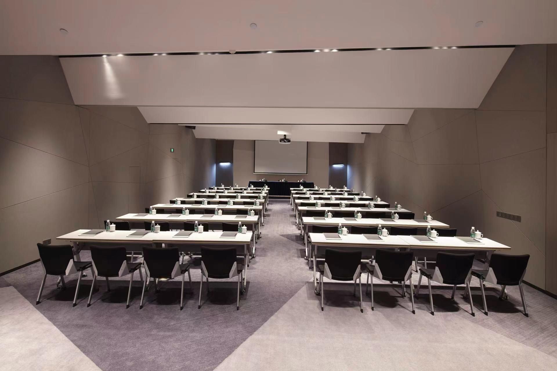 A conference room with rows of tables and chairs, a projector screen, and a stage at the front.