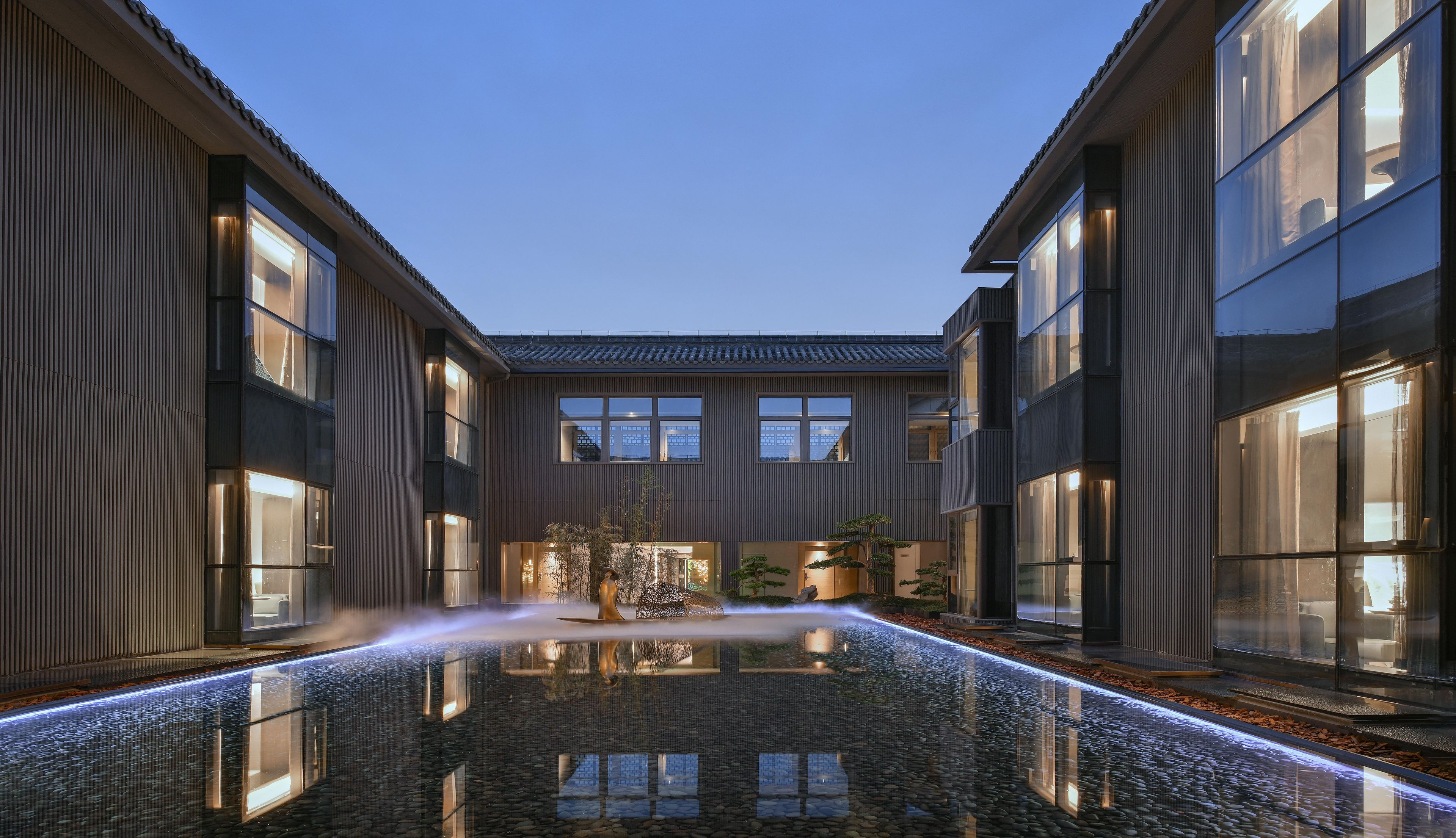 Un edificio moderno con una fuente de agua reflectante en el patio, grandes ventanales y un tejado tradicional de tejas.