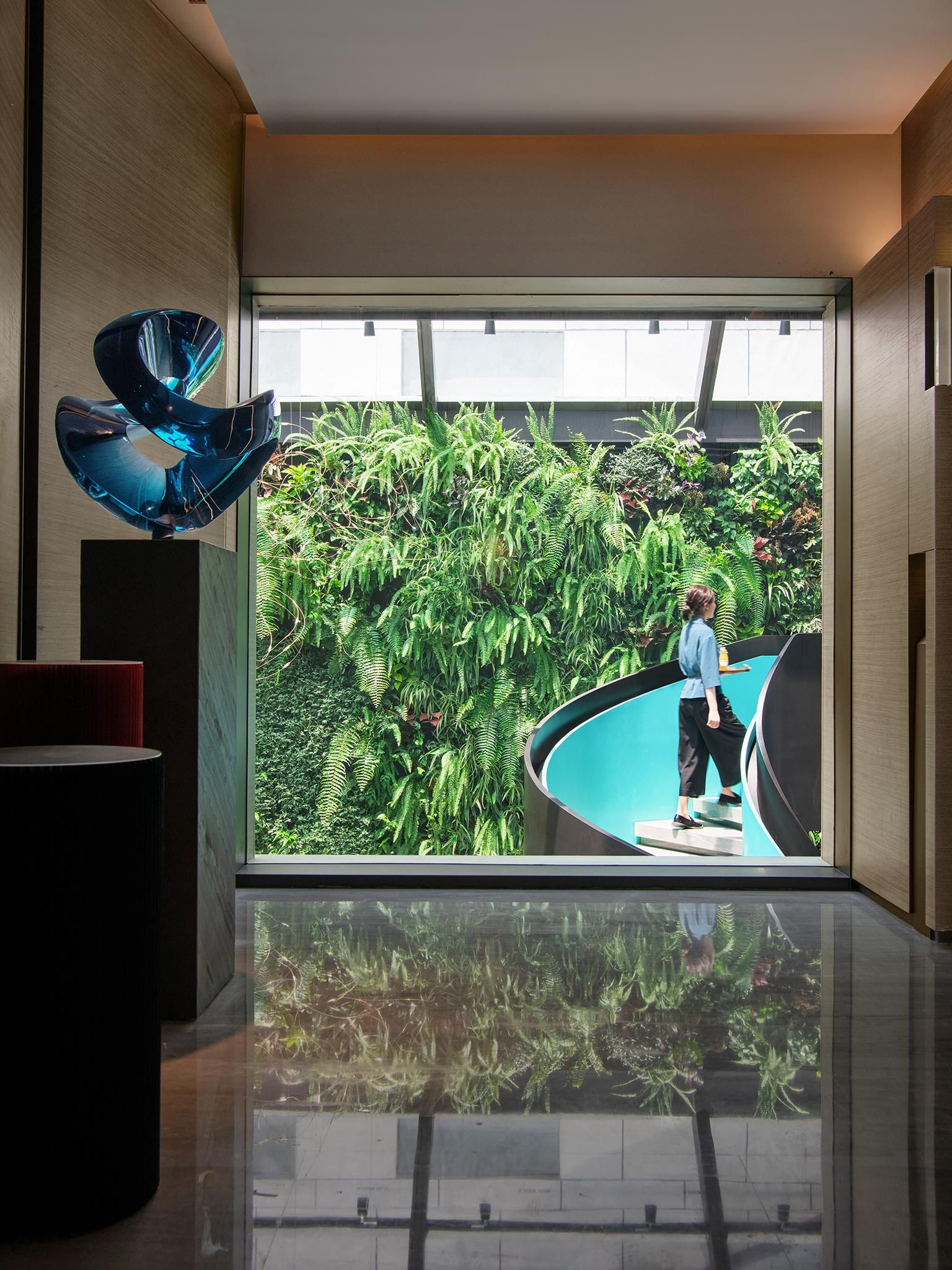 A modern interior with a reflective floor, a glass wall with a view of a lush indoor garden, and a unique blue sculpture.