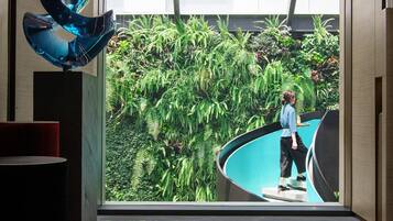 A modern interior with a reflective floor, a glass wall with a view of a lush indoor garden, and a unique blue sculpture.