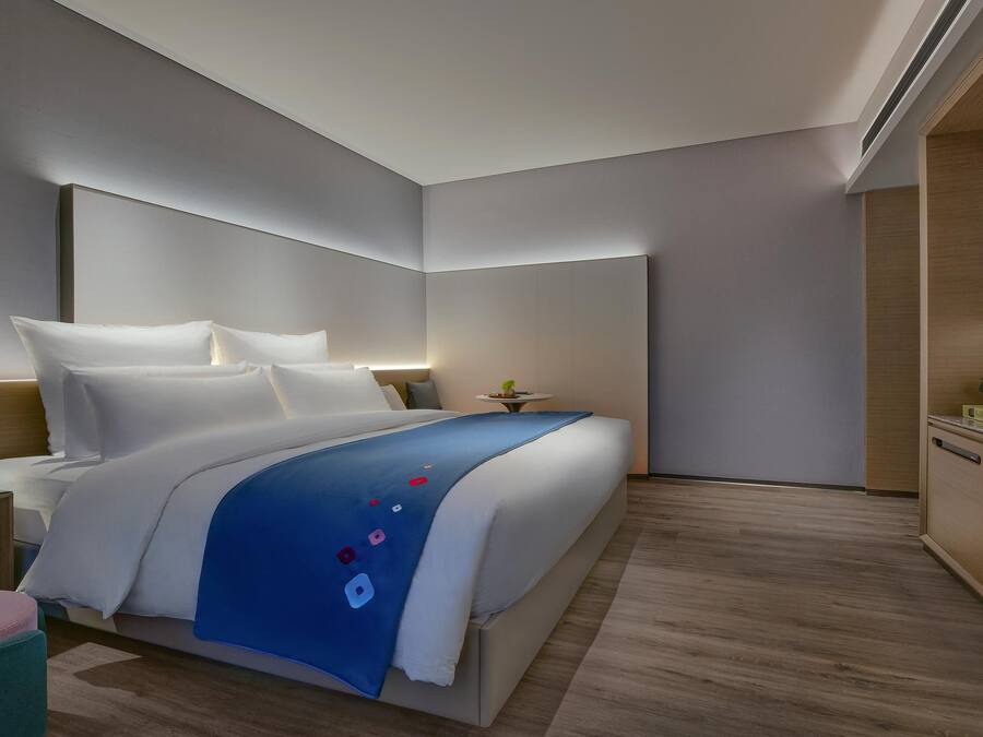 A modern hotel room with a large bed, a bedside table with a lamp, and a small plant.