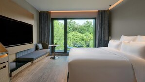 A modern hotel room with a large bed, a flat-screen TV mounted on the wall, a small table with a plant, and a balcony with a view of greenery. - Blossom Hill Hotel Yangzhou Slender West Lake (Yangzhou)