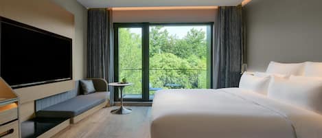 Une chambre d’hôtel moderne dotée d’un grand lit, d’un téléviseur à écran plat fixé au mur, d’une petite table avec une plante et d’un balcon offrant une vue sur la verdure.