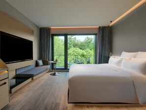 A modern hotel room with a large bed, a flat-screen TV mounted on the wall, a small table with a plant, and a balcony with a view of greenery. - Blossom Hill Hotel Yangzhou Slender West Lake (Yangzhou)
