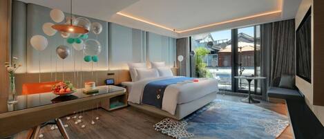 A modern hotel room with a large bed, a desk, a dining table, and a view of the outside.